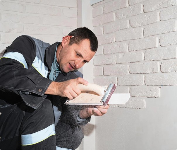 Handyman sanding and smoothing a white brick wall with a sanding block.