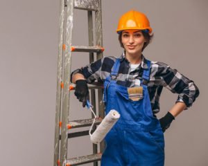 Female painter in overalls and hard hat holding a paint roller beside a ladder.
